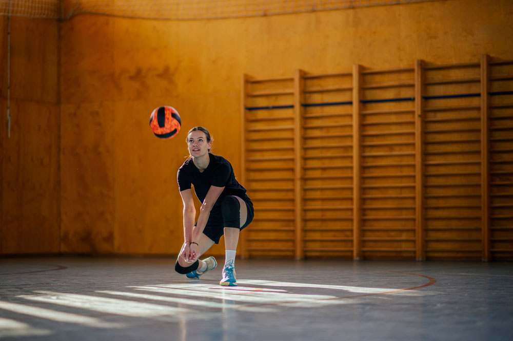 Portrait of a young female volleyball player kneeling during a match or a training while bumping a ball at indoor court. High school volleyball tournament or practice. Copy space.