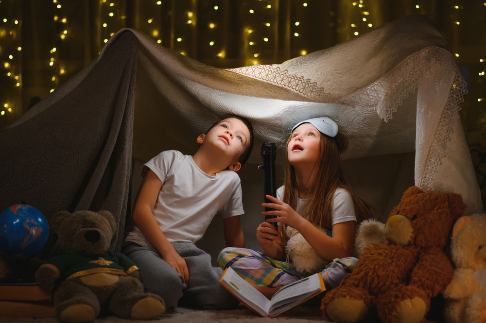 two children playing inside with a makeshift tent and flashlight